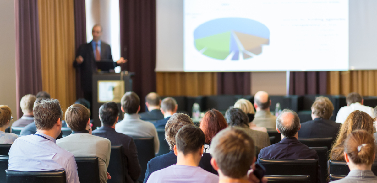 A man in the blurred-out background giving a presentation with a pie chart displayed while attendants watch and take notes in the foreground.