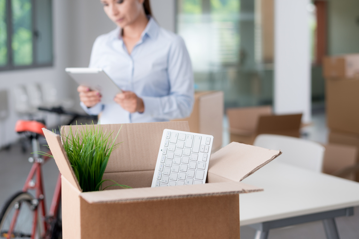A box containing a keyboard and small plant in the foreground, with a woman in the background looking at a tablet.