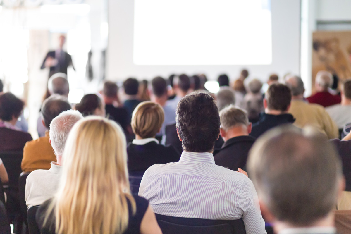 Man speaking to convention space full of people with focus on a business man's back near the last row.