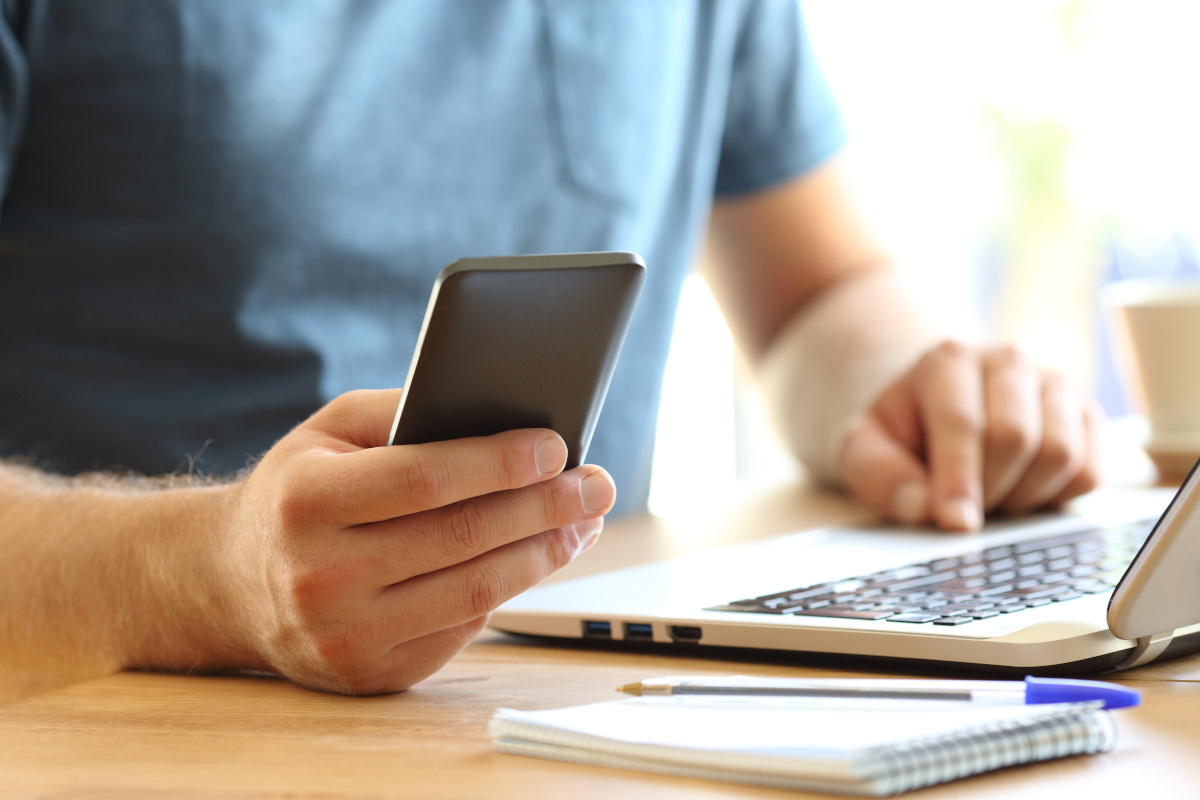 Close up of a man sitting in front of a laptop and holding his mobile phone in his right hand.