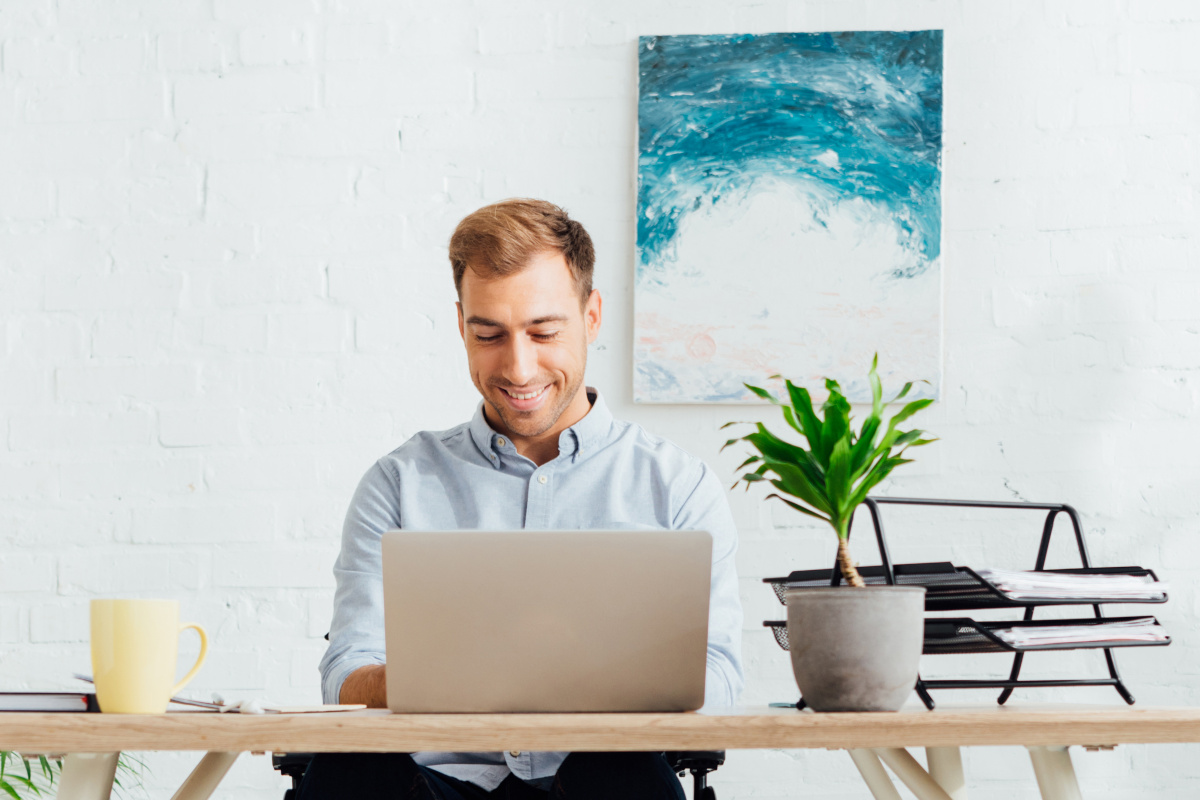 A man smiling while working on his laptop at his desk in his home office